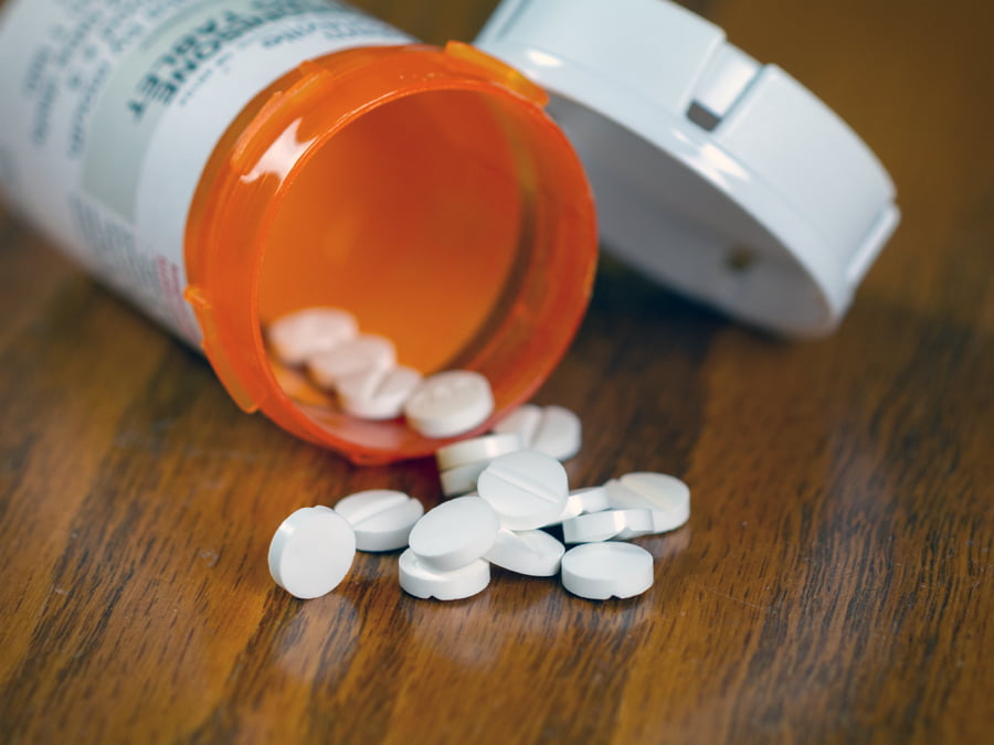 An open pill container is tipped on its side with individual pills spilling out onto a wooden table