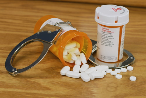Wooden table with handcuffs on it next to an open bottle of pills symbolizing drug laws. The drugs are spilled all over the table.