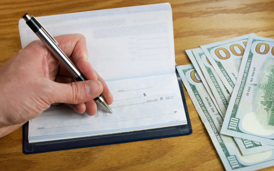 A checkbook is shown with cash off to the side on a wooden table. An individual's hand is seen writing on the check using a zebra mechanical pen.