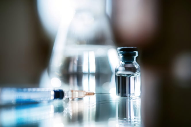 A hypodermic needle is shown in the foreground with a liquid drug vile on a table, with a chemistry flask in the background