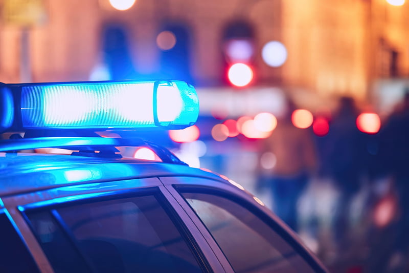 The top of a police car is shown with roof mounted lights illuminated, showing people in the background