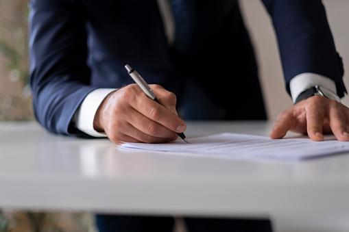 Featured: Male attorney in suit standing at desk writing on a document- Hiring a Private Criminal Defense Attorney